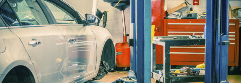 A white car in a car repair shop with its bonnet open, parked near a blue hydraulic lift, red tool chest, and various equipment and tools in the background.