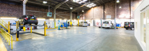 Spacious auto garage with cars parked and one car lifted on a yellow hydraulic ramp, tools and equipment along the walls, and a high ceiling with exposed brick and skylights letting in natural light.